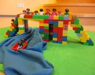 Bunte Bausteine bilden eine Brücke, auf der Spielzeugfiguren stehen. Darunter deutet eine blaue Decke einen Fluss an, auf dem ein Spielzeugboot liegt.