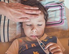 Ein Kind liegt im Bett mit einem Fieberthermometer im Mund und eine Hand befühlt die Stirn.