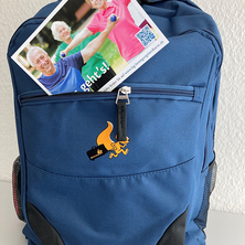 Ein blauer Rucksacktrolley, in der Außentasche steckt eine Postkarte.