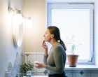 Eine Frau steht im Badezimmer vor einem Spiegel und cremt sich das Gesicht ein.