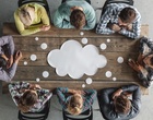 Von oben betrachtet, sitzen acht Personen um einen Tisch herum und schauen auf die Tischplatte, auf der eine Gedankenwolke ist, Symbolbild für Teammeeting und Ideenfindung.