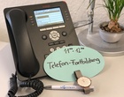 Auf einem Schreibtisch steht ein Telefonapparat mit einer Zettelnotiz "11:00 - 12:00 Telefonfortbildung". Daneben liegen eine Armbanduhr und ein Kugelschreiber.