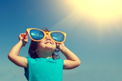Ein Kind hält sich eine übergroße Sonnenbrille vor das Gesicht und schaut in den Himmel.